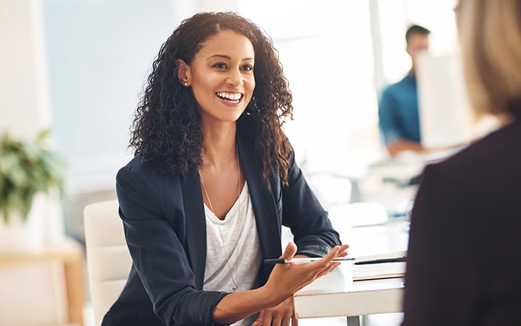 A business professional woman seated at a desk and actively gesturing with her hands, possibly explaining or discussing something to the person in front of her.