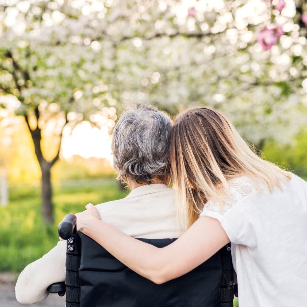 A young woman with blonde hair wearing a white dress, embracing an older woman in a wheelchair with grey hair.