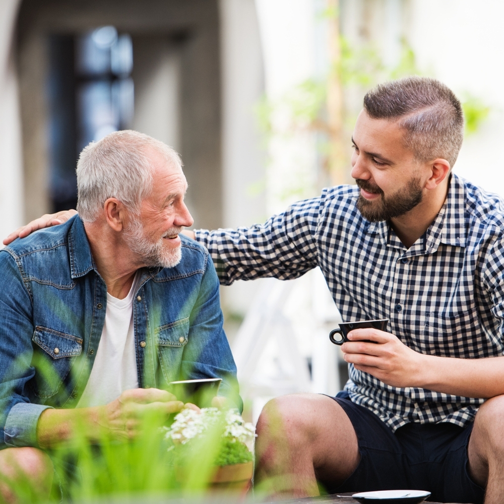 Two men sitting outdoors and engaged in conversation, one older with white hair and a beard, wearing a denim jacket, and the other younger in a black and white checkered shirt.
