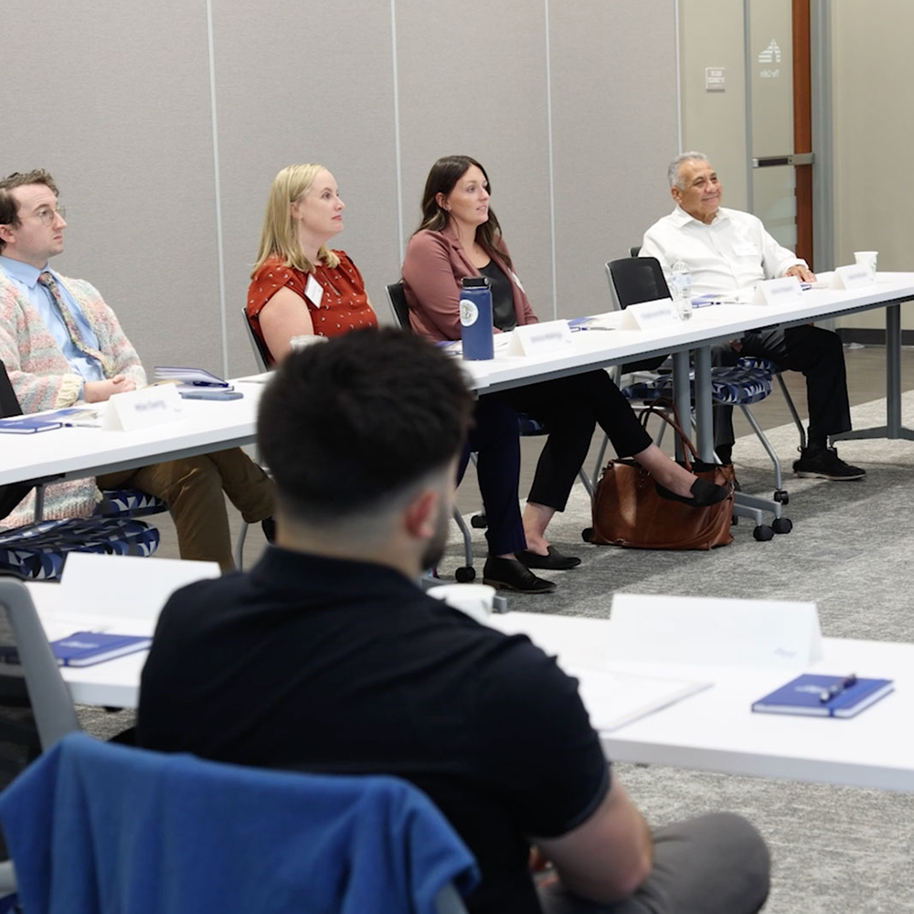 A group of professionals sits at a conference table, listening to a speaker present